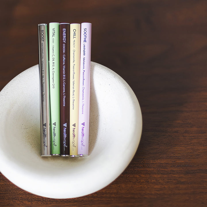 Five HealthVape pens in assorted flavors displayed in a white bowl on wooden surface