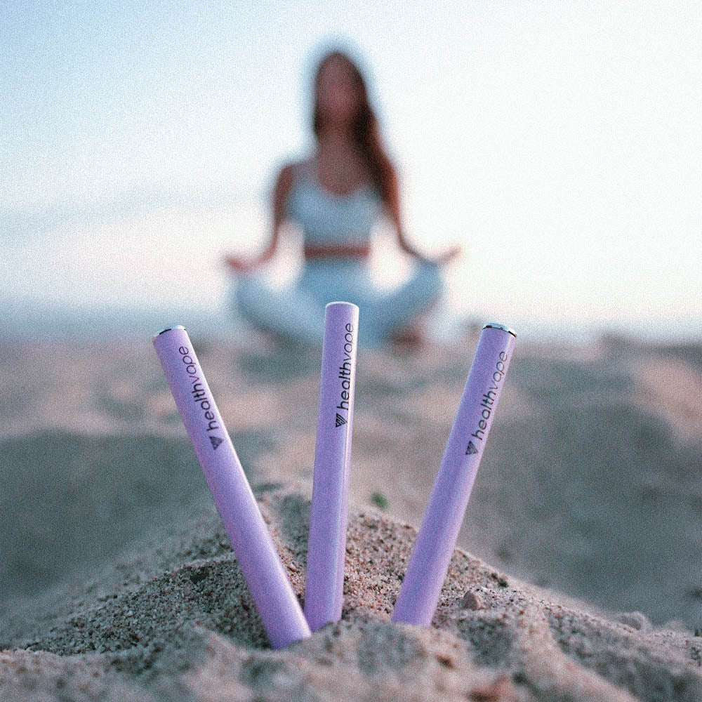 Three HealthVape Chill Lavender disposable vape pens standing in sand with blurred yoga figure