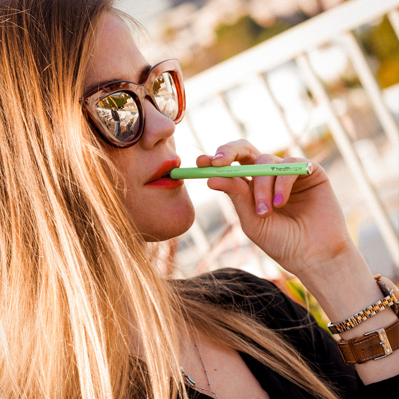 Woman wearing sunglasses using a green HealthVape ENERGY disposable inhaler outdoors with a blurred background.